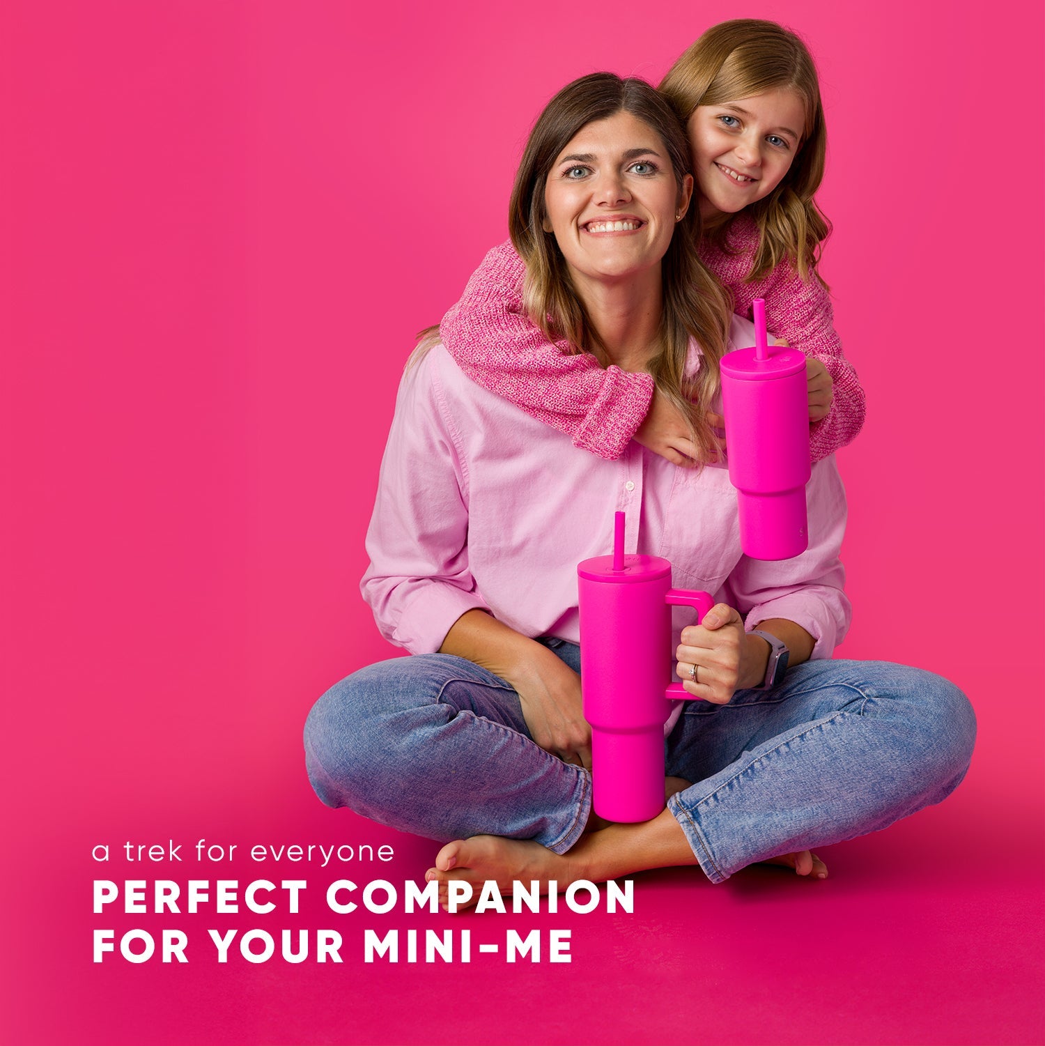 Adult woman and young child seated together on hot pink background, both wearing matching magenta knit sweaters and blue jeans, holding bright fuchsia Simple Modern Trek tumblers with straws. Text reads "a trek for everyone"