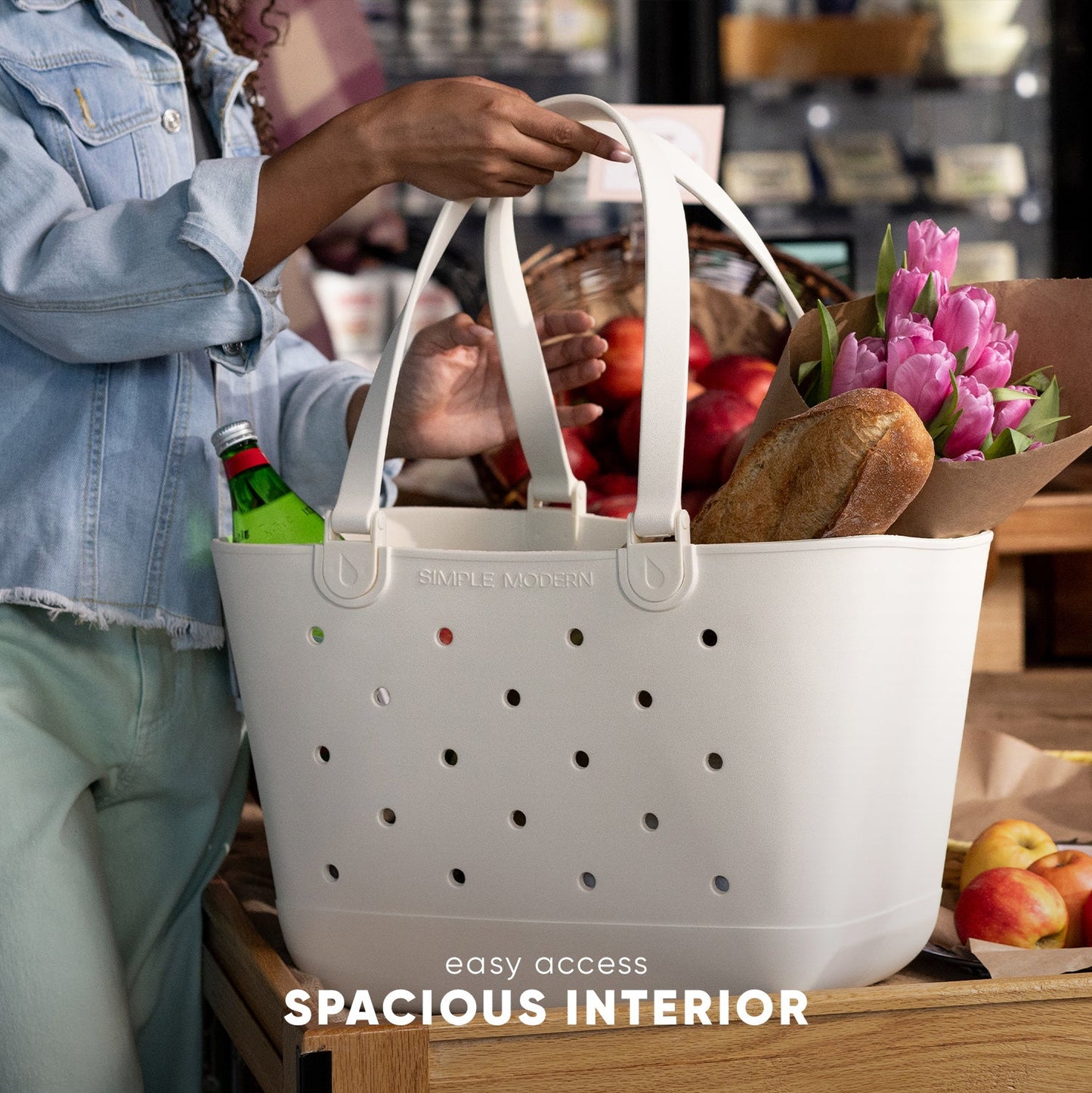 White Simple Modern tote bag with perforated ventilation held by person in light blue denim, filled with fresh tulips, bread, produce, and groceries on wooden table in market setting.