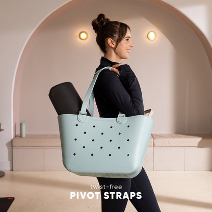 Woman wearing black top carrying light mint-green Simple Modern tote bag with perforated pattern and twist-free pivot straps in modern studio setting.