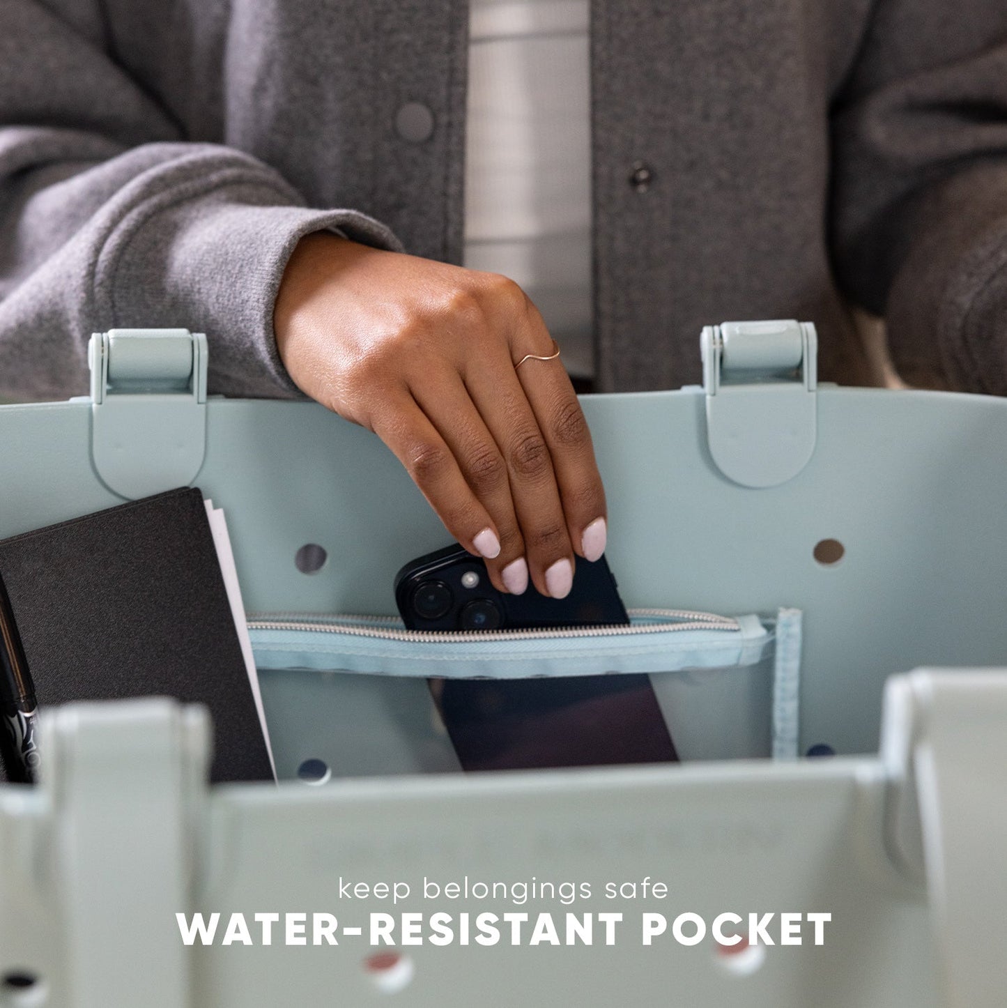 Sage green tote bag with person placing black smartphone into water-resistant zippered pocket. Text reads "keep belongings safe" and "WATER-RESISTANT POCKET." Studio lifestyle product photo.
