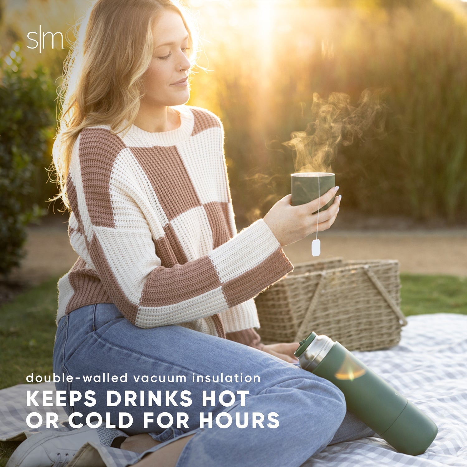 Woman wearing tan and cream checkered sweater and blue jeans holding a double-walled vacuum insulated beverage container in golden hour sunlight.